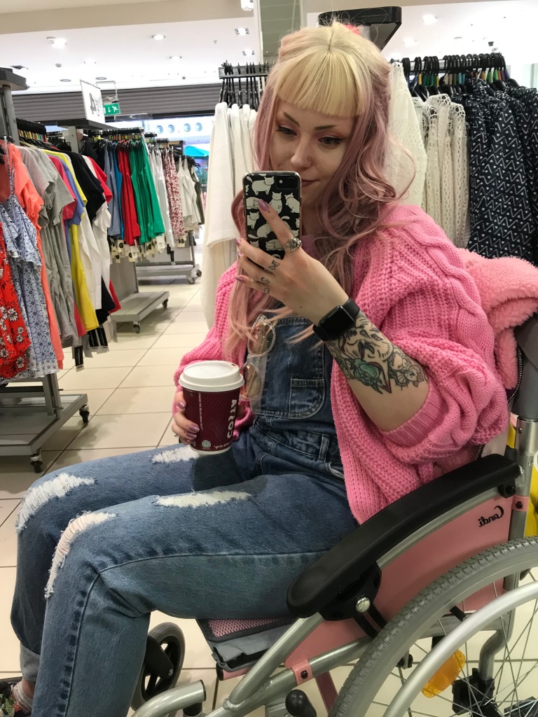 Pixie sits in her wheelchair, holding a coffee cup. She is taking a selfie in a shop mirror. She is wearing a long pink, purple and blonde wig, with pink makeup. She is also wearing a pink knitted cardigan and blue denim dungarees.