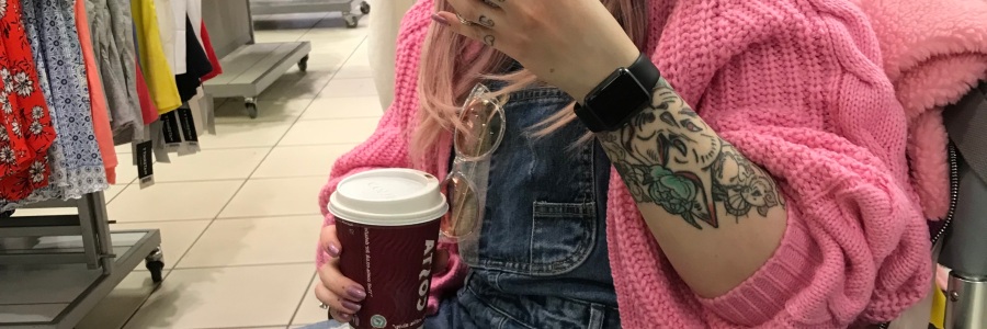 Pixie sits in her wheelchair, holding a coffee cup. She is taking a selfie in a shop mirror