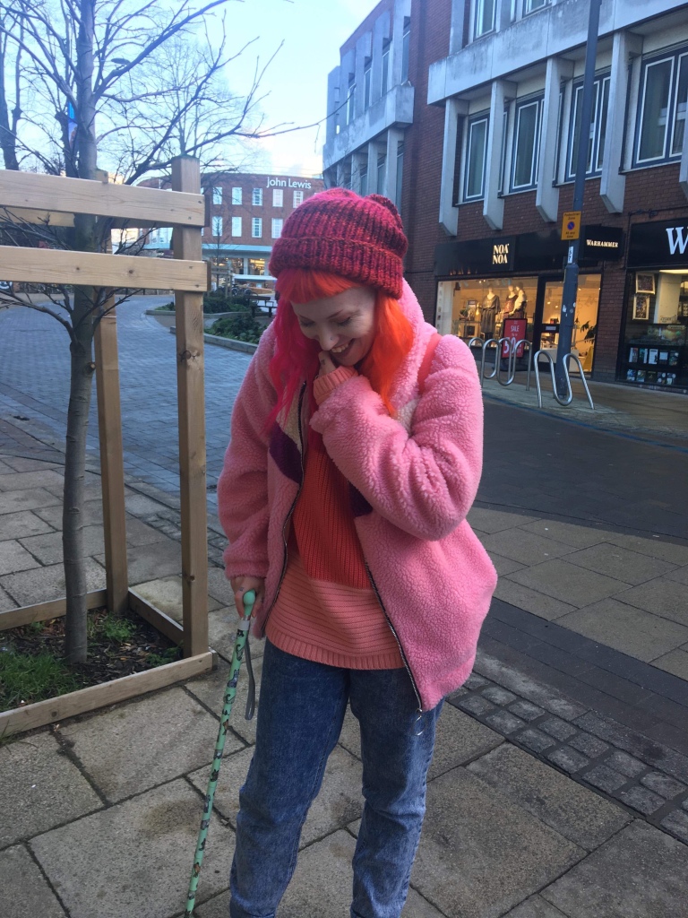 Pixie is standing in a wintry town scene, leaning on a mint green walking stick decorated with images of cartoon cats. She smiles shyly and looks toward the ground instead of the camera, holding one hand to her face. In this image she has long pink and orange half-and-half hair and is wearing a pink fluffy jacket, a red knitted bobble hat, a knitted jumper in varying shades of pink, and blue jeans.