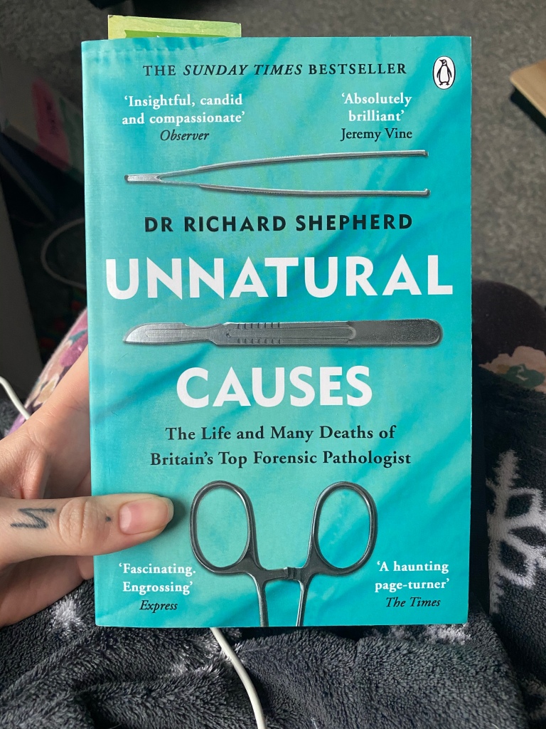 Pixie is holding a book, titled "Unnatural Causes" by Doctor Richard Shepherd. The cover is light blue, and has images of various surgical instruments lined up across the front. There are quotes from readers such as "absolutely brilliant" and "a haunting page-tuner" dotted around.